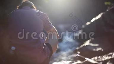在阳光明媚的日子里，放松的年轻人用智能手机坐在瀑布旁的岩石上拍照。 1920x1080